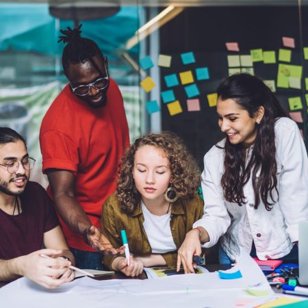 Group of multiracial casual women and men gathering at table with paper poster and sharing ideas for new creative project