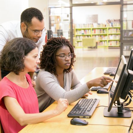Trainer helping students in computer class. Man and women sitting and standing at desk, using desktop, pointing at monitor, asking, talking. Education concept