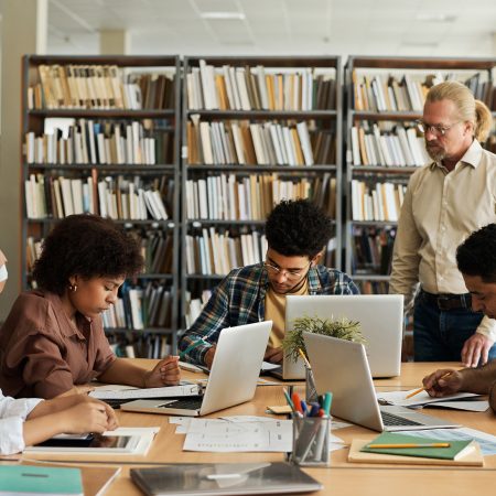 Group of foreign students learning language sitting at table with computers and textbooks together with teacher explaining material